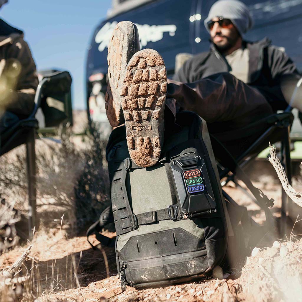 Person sitting on a chair with their feet up on a backpack with a Thyrm CellVault 5M attached. Person is in a rugged outdoor setting, and a vehicle is in the background.
