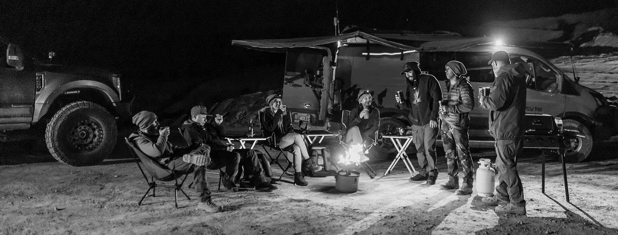 Group of people camping outdoors at night with vehicles and a fire pit.