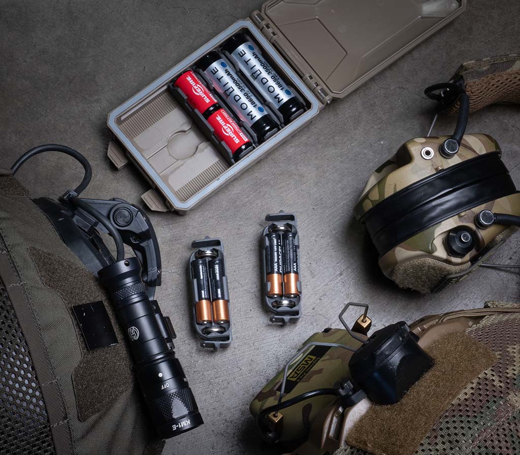 Assorted tactical gear, including a Thyrm CellVault-5M, flashlight, batteries, and ear protection on a textured surface.