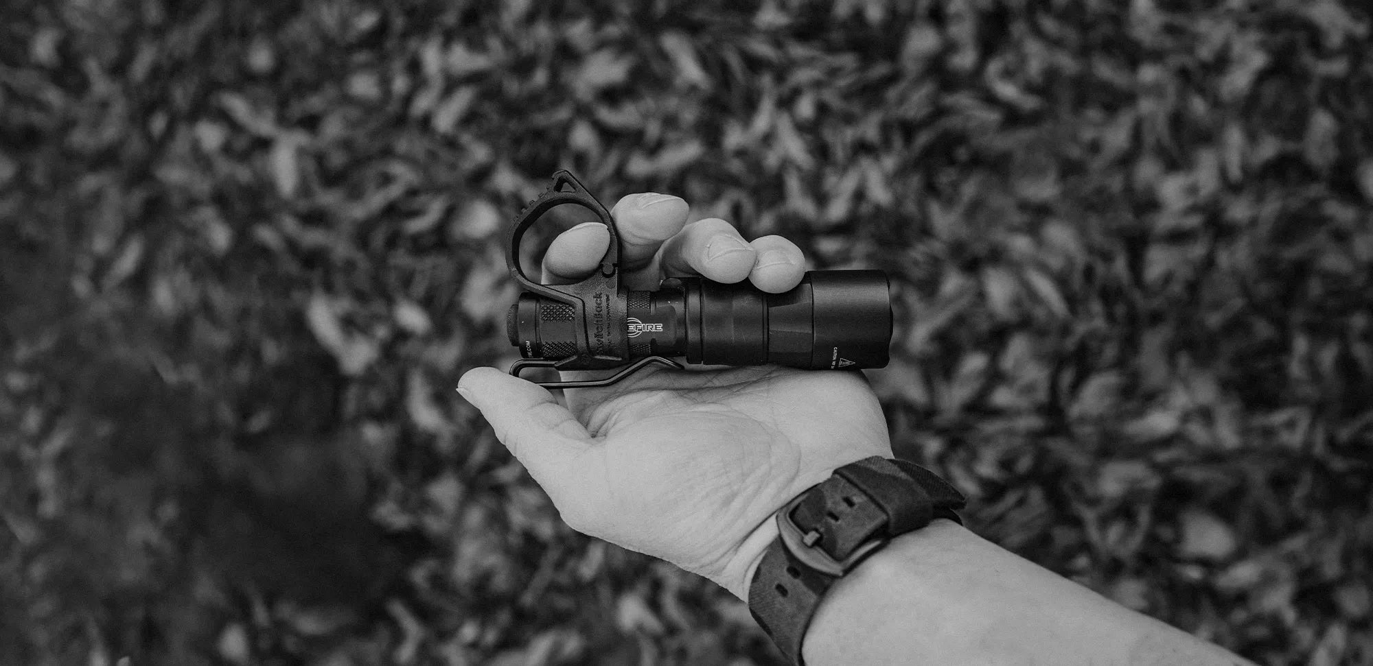 Hand holding a Surefire flashlight with a Thyrm SwitchBack against a blurred natural background.