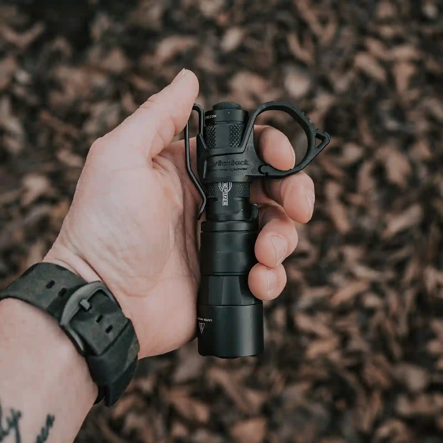 Hand holding a black Surefire flashlight with a Thyrm SwitchBack against a blurred natural background