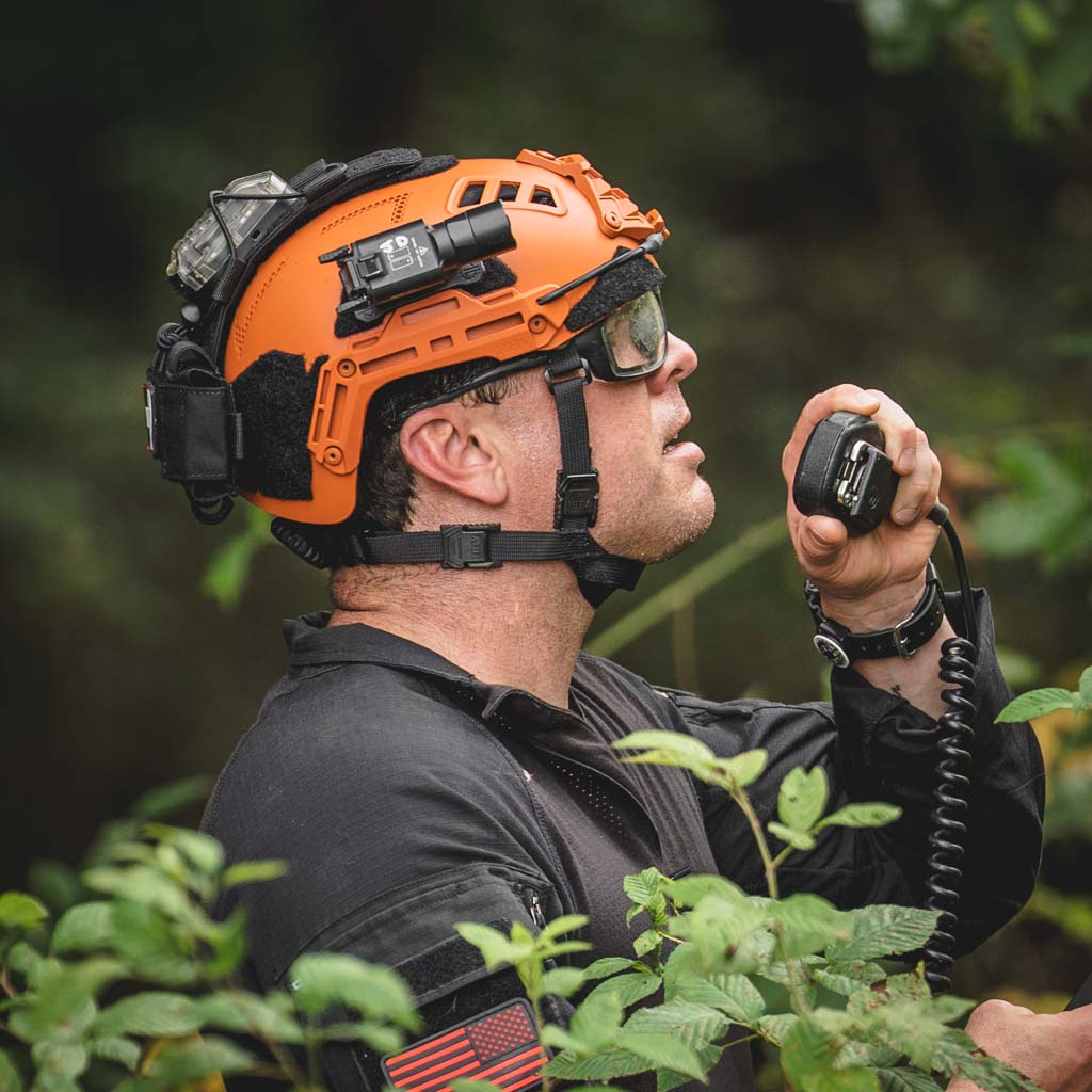 Person wearing an orange helmet with a Thyrm VariArc attached, holding a radio in a forest setting.