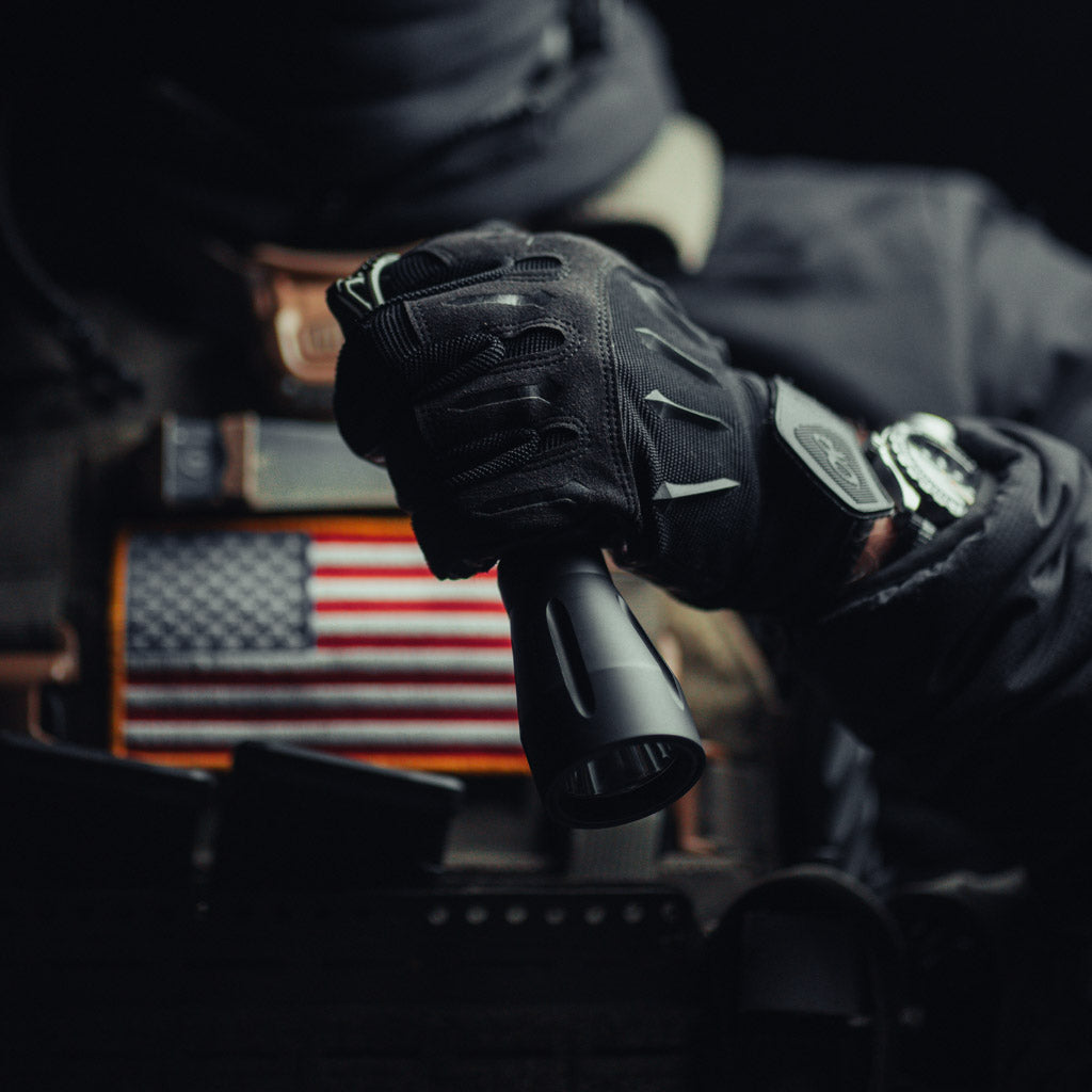 Person wearing tactical gloves holding a Modlite flashlight with a Thyrm Switchback with an American flag patch in the background