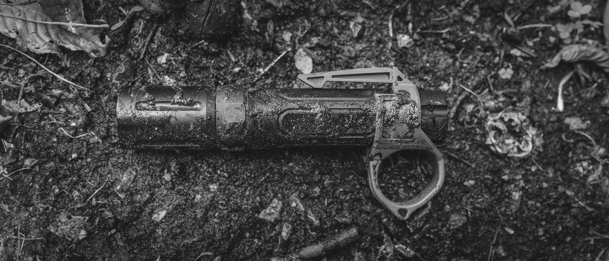 Muddy Modlite flashlight with a Thyrm Switchback, lying on muddy ground surrounded by leaves and debris.