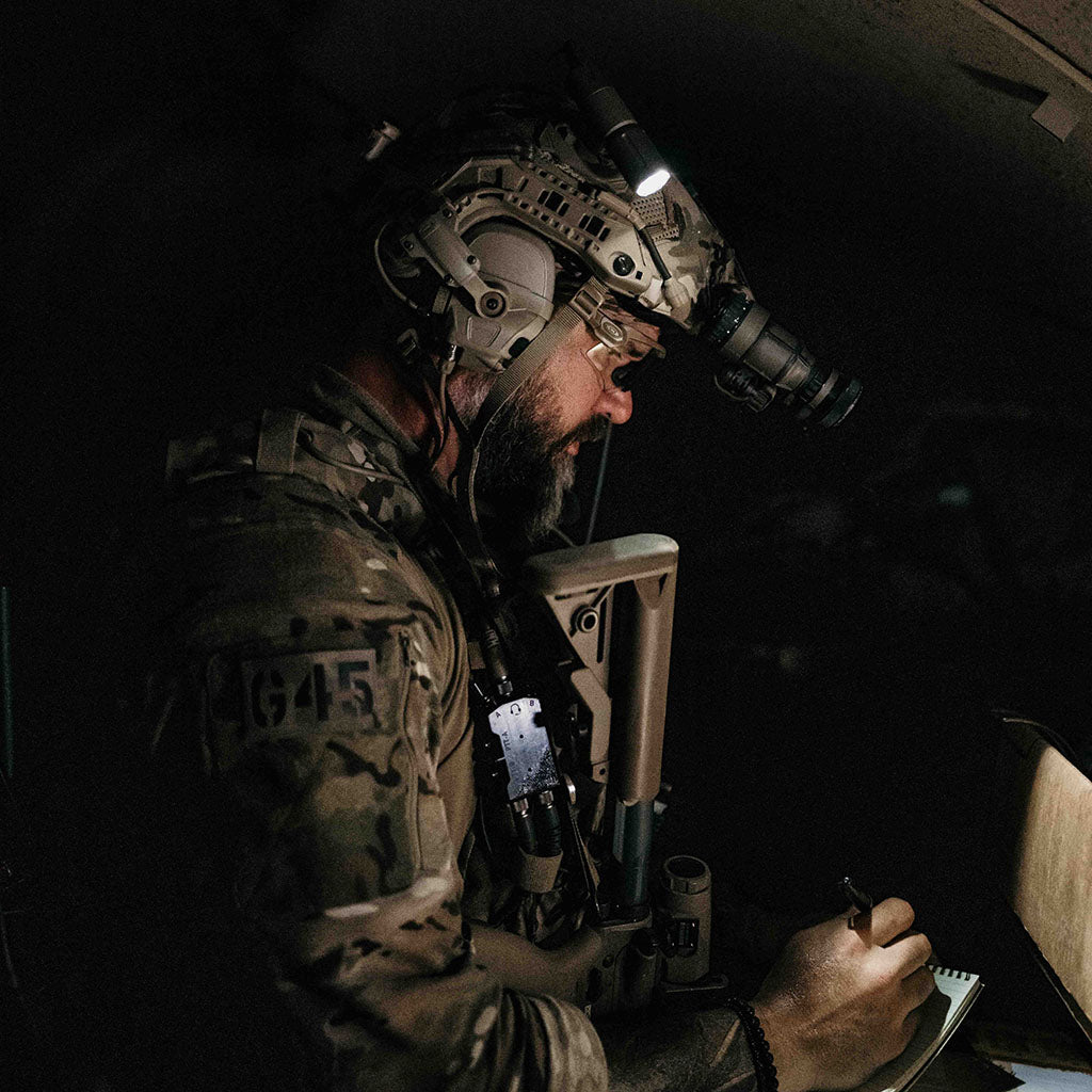 Person in military gear with a Thyrm VariArc and night vision goggles in a dark setting, planning on a tablet of paper.