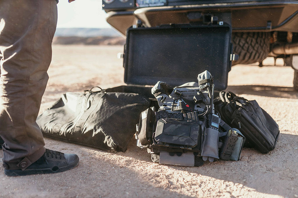 Person standing next to a black toolbox and equipment on a dirt road.  He has his chest rig configured with mounted gear including a DarkVault Comms Critical Gear Case in MultiCam Black.